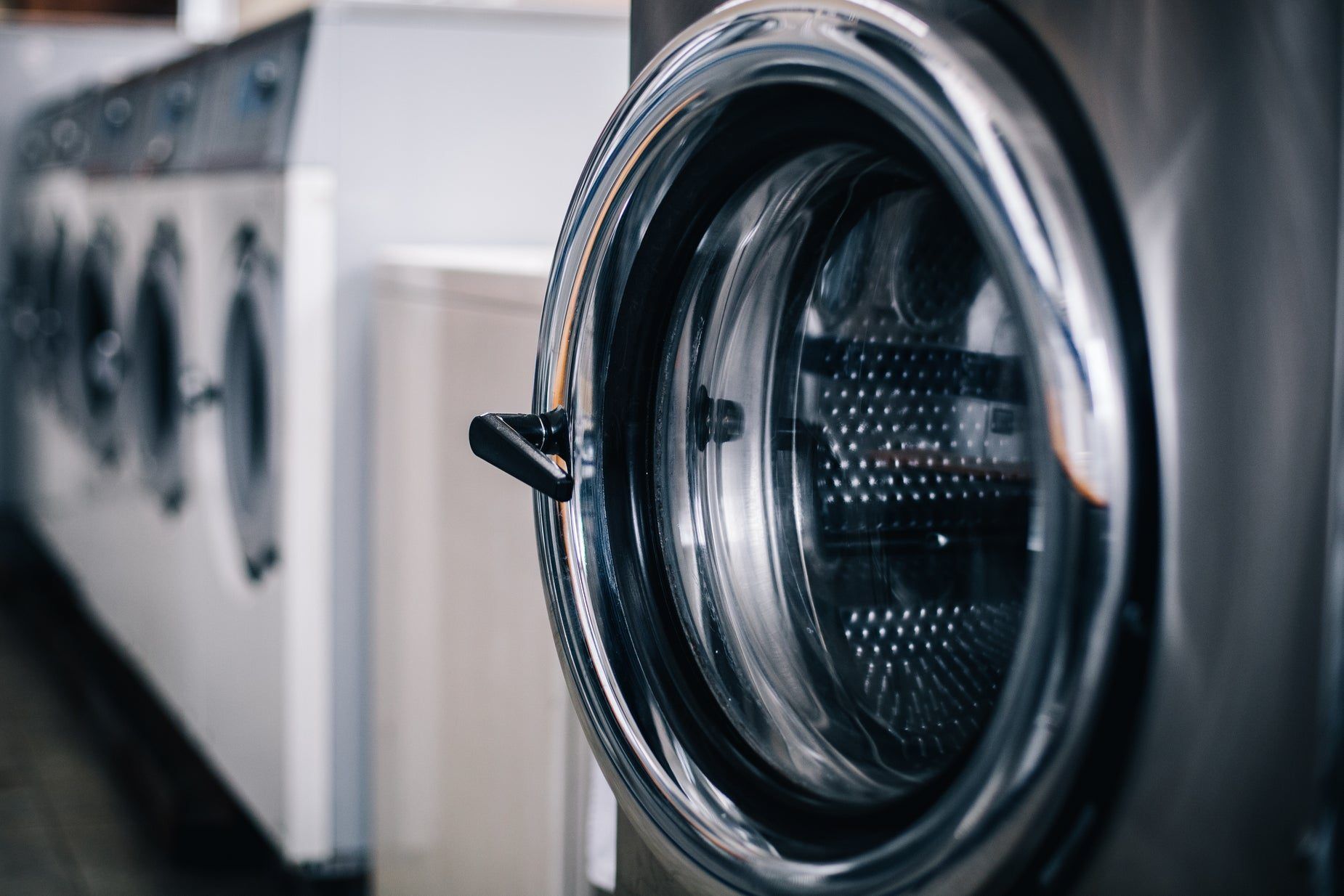 Open glass door of a stainless front-load washer showing the perforated drum, more machines blurred behind