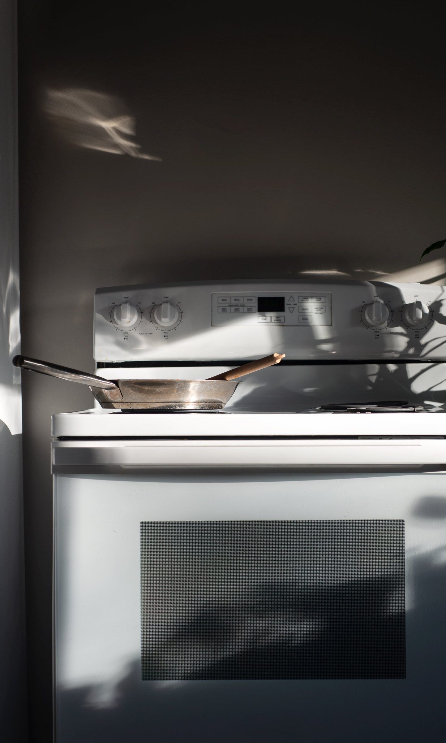 White electric range with a frying pan on the cooktop, raked by window light and leaf shadows